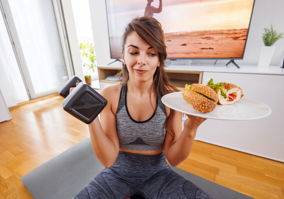 Woman in sportswear holding a dumbbell in one hand and sandwich on a plate in the other one - choice between healthy lifestyle and workout and fast food